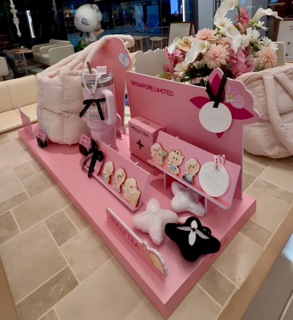 The “Singapore Limited” merchandise island at Raffles Place — cloud totes, pearl-strapped tumblers and blind-box magnets, staged like a beauty counter. 